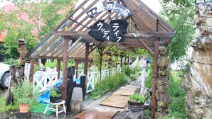 A wooden gazebo with a sign, surrounded by potted plants and a wooden pathway. - Woody Life (Kamifurano)