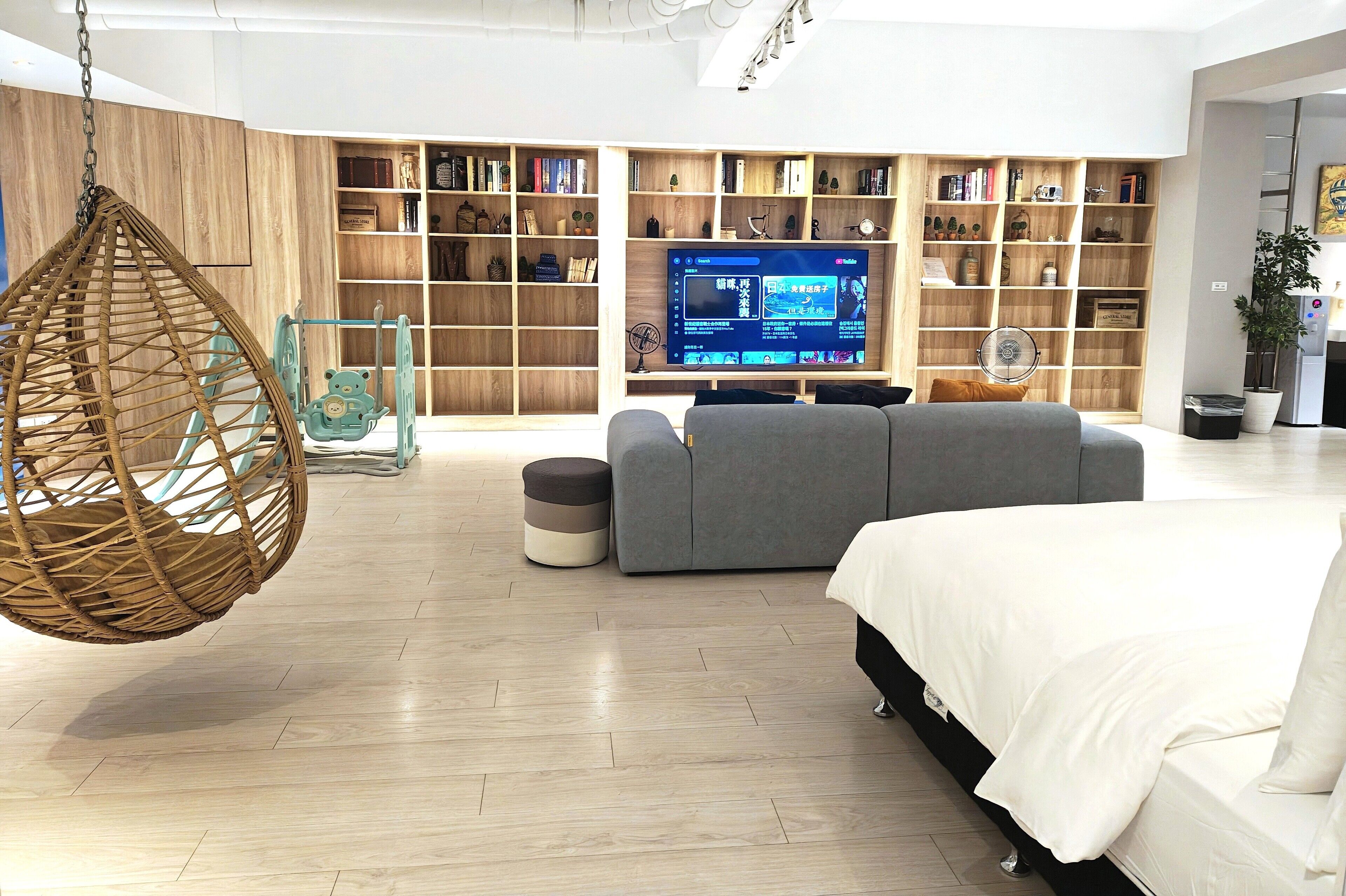 A modern living room with a large bookshelf, a hanging chair, a grey sofa, and a bed with white bedding.