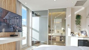 Egy modern szállodai szoba, amelyben egy nagy ágy, a falra szerelt televízió, egy fürdőszobába nyíló, toló üvegajtó, valamint egy kis íróasztal és egy kávéfőző található.