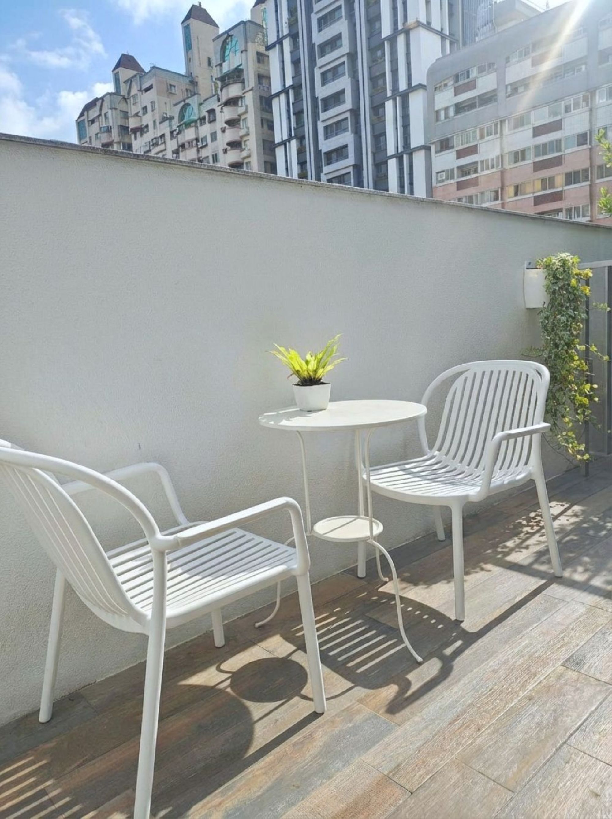 Un pequeño espacio al aire libre con dos sillas blancas y una mesa redonda, una planta en maceta, y vista a un paisaje urbano con edificios de varios pisos.