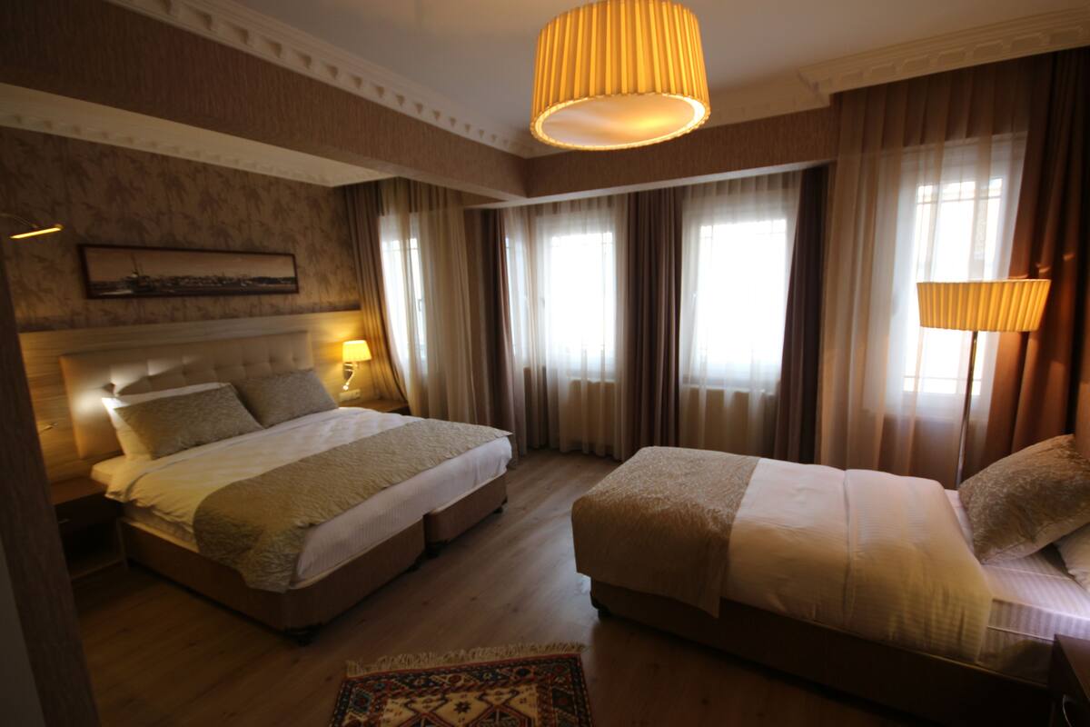 A hotel room with two beds, a large window with curtains, a mounted artwork on the wall, and a patterned rug on the floor.