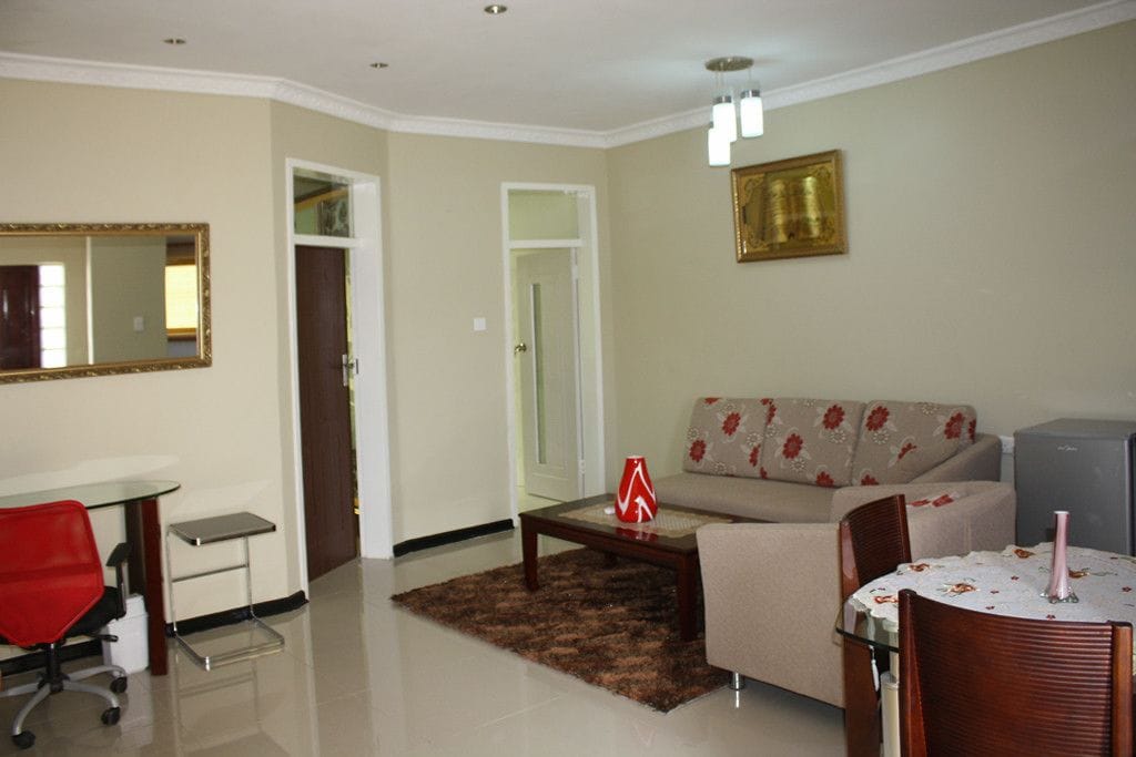 A living room with a sofa, a coffee table, a dining table with chairs, a red office chair, a small table with a lamp, and a mirror on the wall.