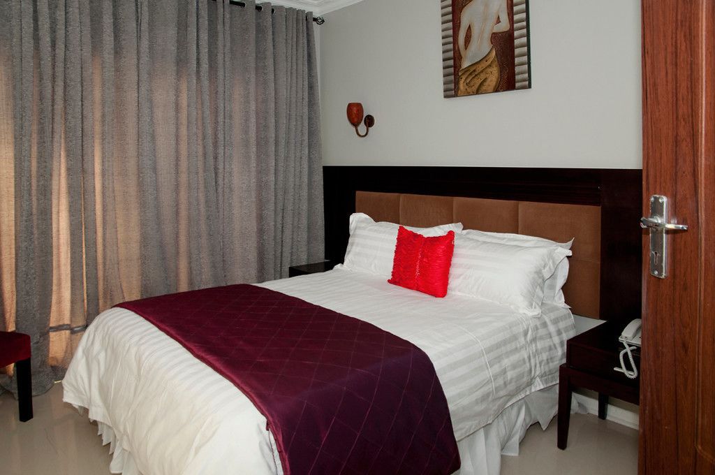 A neatly made bed with white linens and a red accent pillow, a wooden nightstand with a telephone, and a painting on the wall.