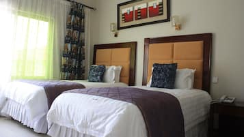 A hotel room with two beds, a large window with sheer curtains, a framed artwork above the beds, and a bedside table with a phone.