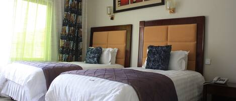 A hotel room with two beds, a large window with sheer curtains, a framed artwork above the beds, and a bedside table with a phone.
