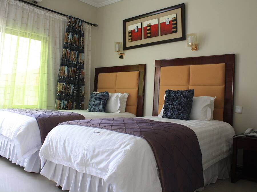 A hotel room with two beds, a large window with sheer curtains, a framed artwork above the beds, and a bedside table with a phone.