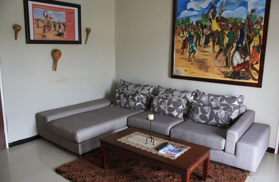 A living room with a grey sectional sofa, patterned pillows, a coffee table with magazines, a framed picture on the wall, and a painting depicting a dance scene.