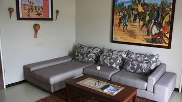A living room with a grey sectional sofa, patterned pillows, a coffee table with magazines, a framed picture on the wall, and a painting depicting a dance scene.