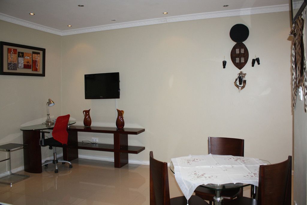 A dining area with a table set for four, a red chair, a desk with a computer, and a flat-screen TV mounted on the wall.