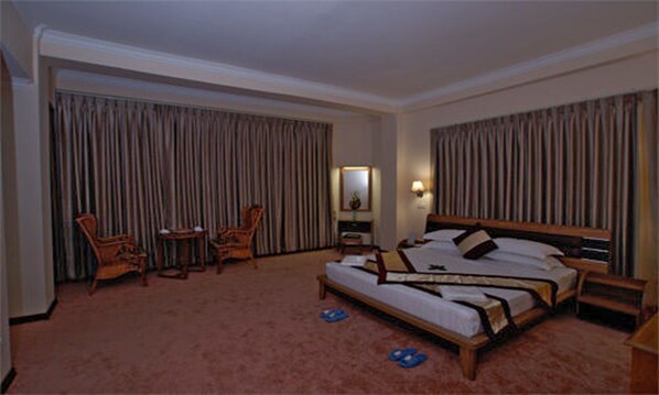 Quarto de hotel com uma cama grande, duas cadeiras, uma mesinha e uma mesa de cabeceira.