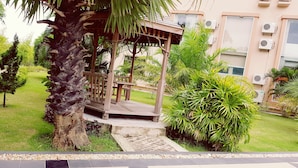 A gazebo with a wooden roof and bench, surrounded by lush greenery and a paved walkway. - Oattara Thiri Hotel (Oattara Thiri Township)