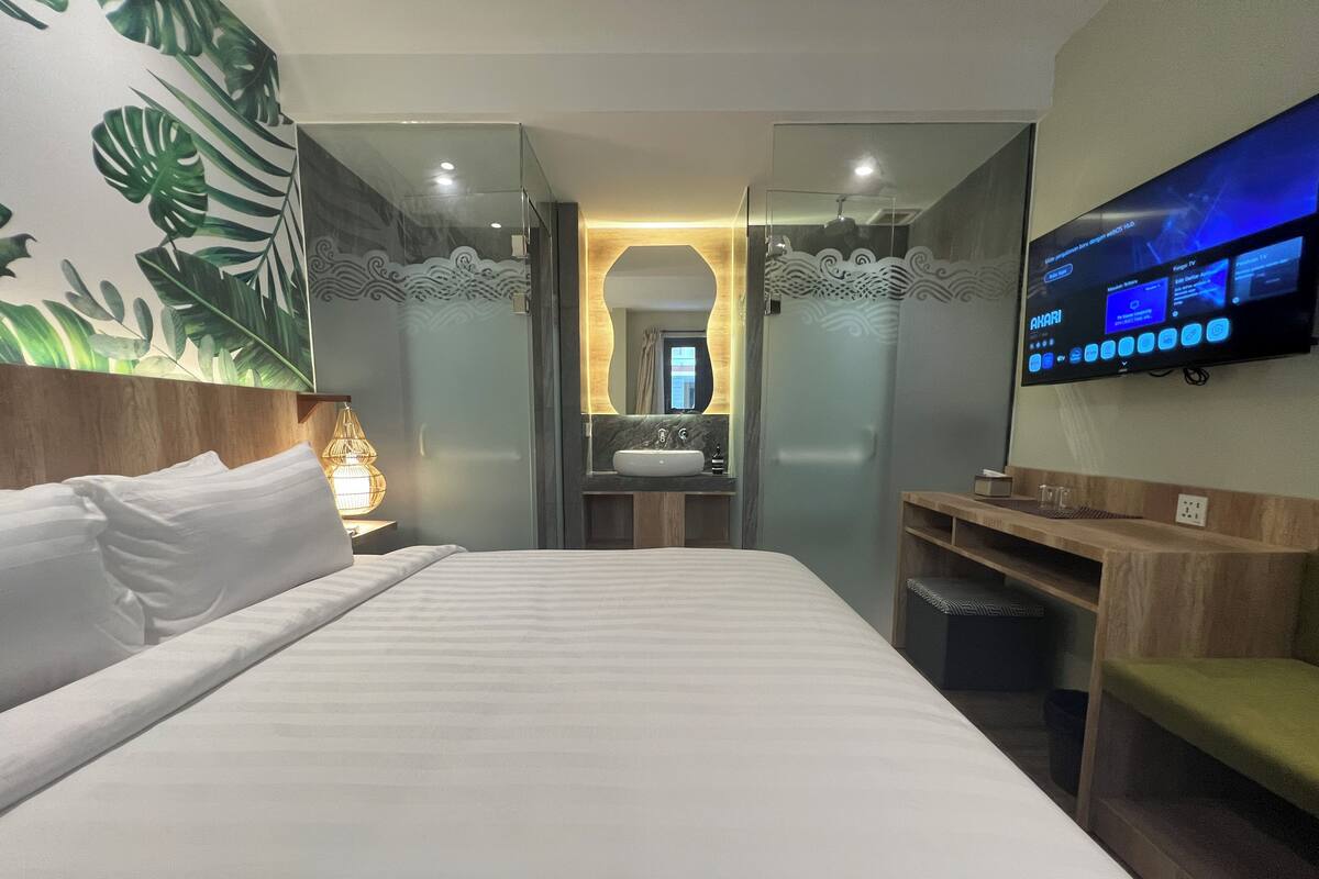 Habitación de hotel con cama, televisor montado en la pared, baño con lavamanos y espejo, y una pared decorativa con motivos vegetales.