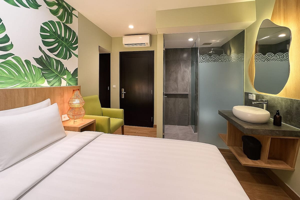 Habitación de hotel con cama, una silla verde, baño con lavamanos y espejo, y ducha.