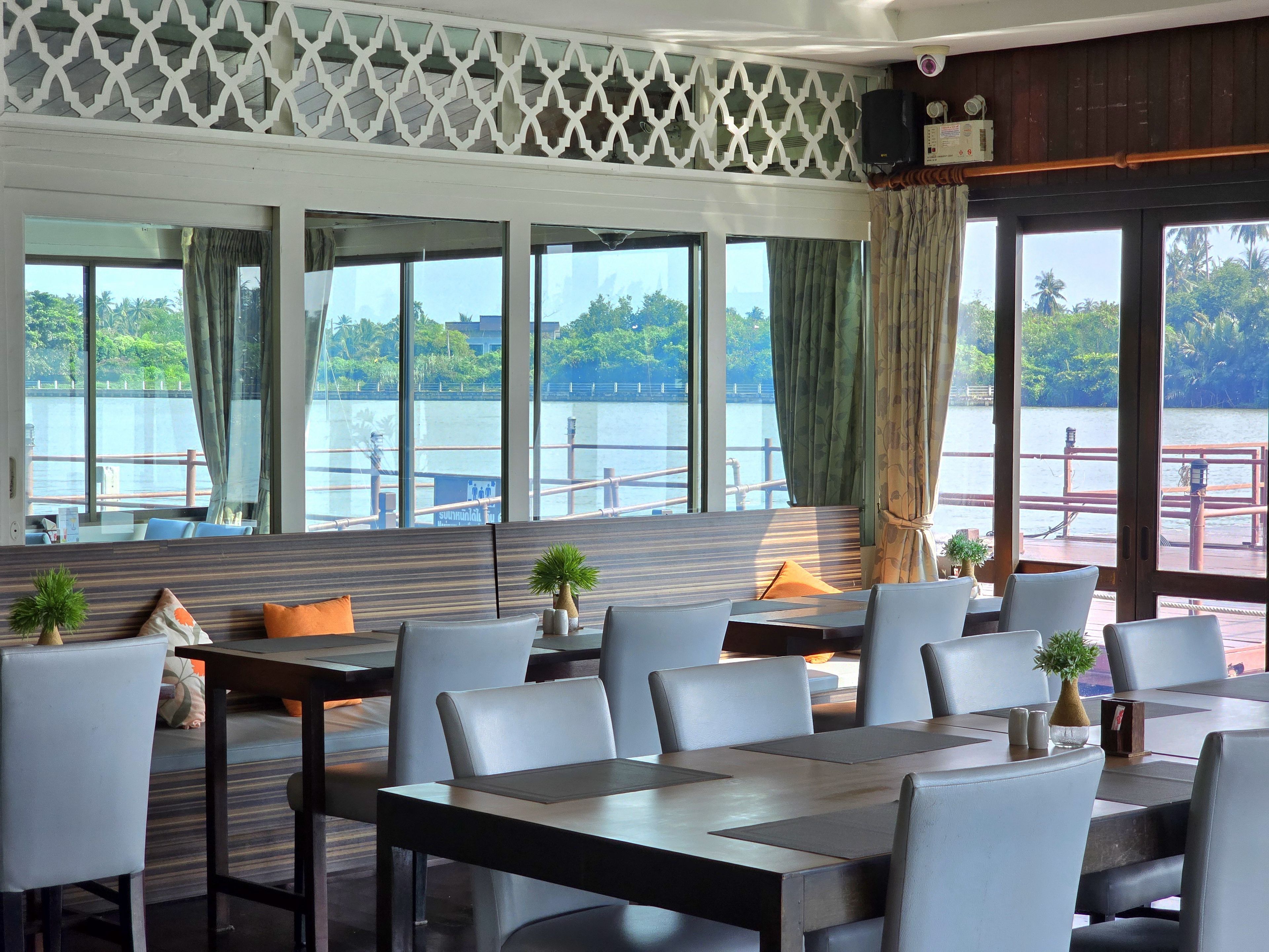 Un restaurante con vistas a un lago, que cuenta con una larga mesa con sillas, un techo estampado y una baranda de madera en el exterior.