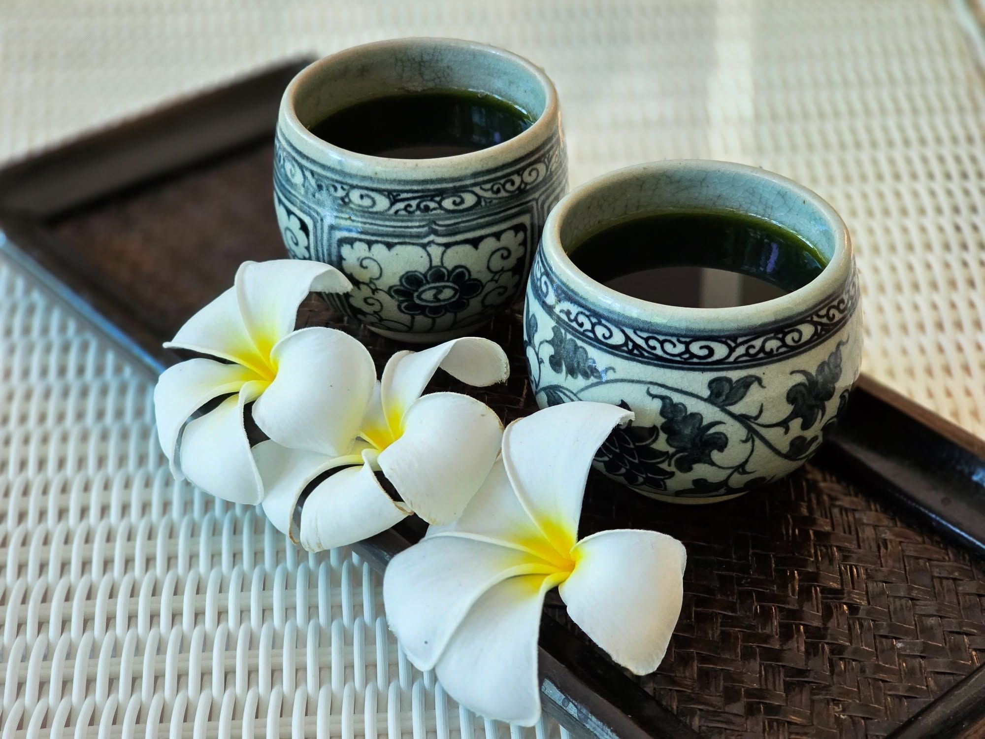Due tazze di ceramica con decorazioni floreali su un vassoio intrecciato, con fiori bianchi.