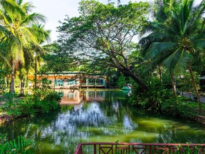 A serene water body with a wooden bridge, lush greenery, and a building with a green roof. - Baan Amphawa Resort & Spa (Amphawa)