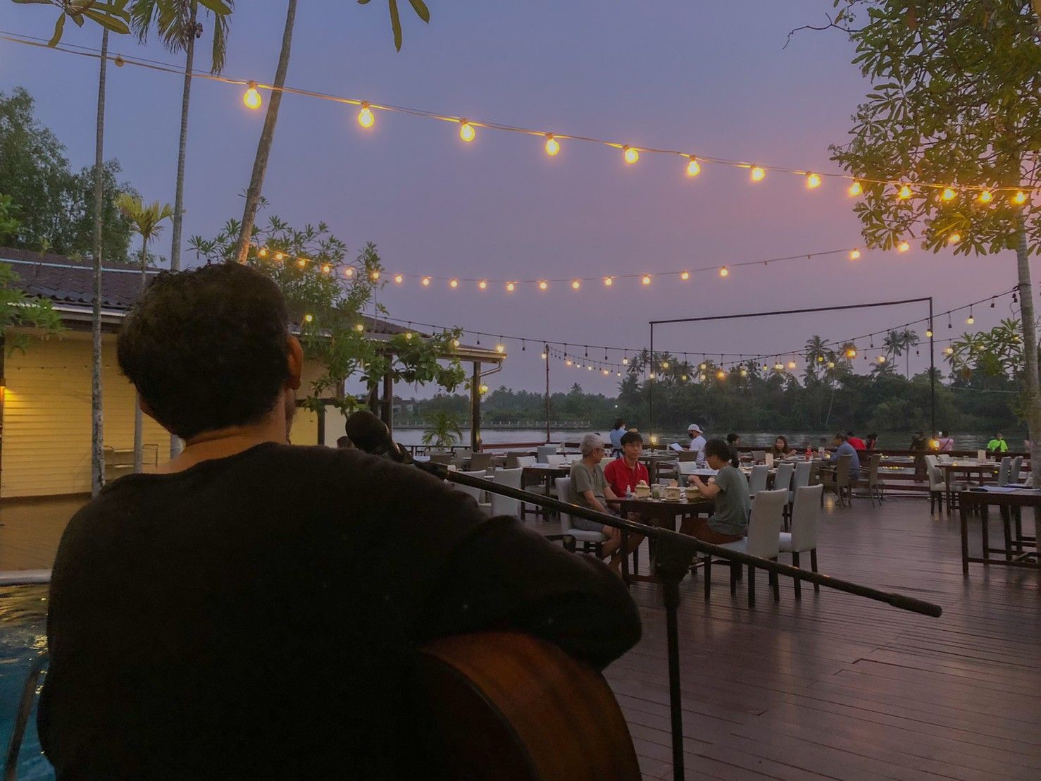 Un espace extérieur aménagé pour les repas, avec des tables et des chaises, des guirlandes lumineuses suspendues et une vue sur l’eau.