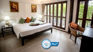 A hotel room with a bed, two bedside tables with lamps, a wicker chair, and large windows with a view of trees. - Baan Amphawa Resort & Spa (Amphawa)
