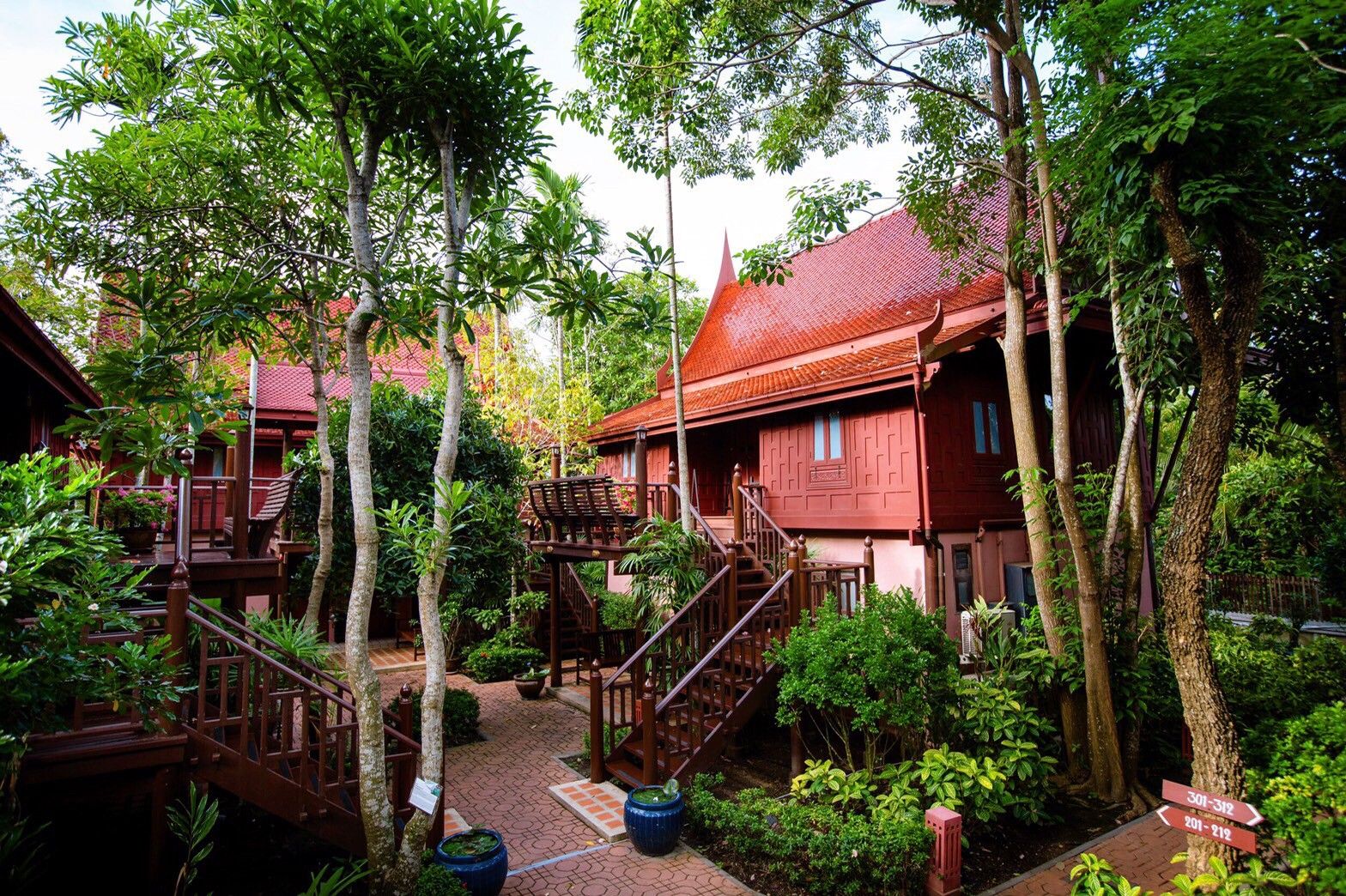 A traditional wooden house with a red roof, surrounded by lush greenery and a wooden staircase leading to the upper floor.