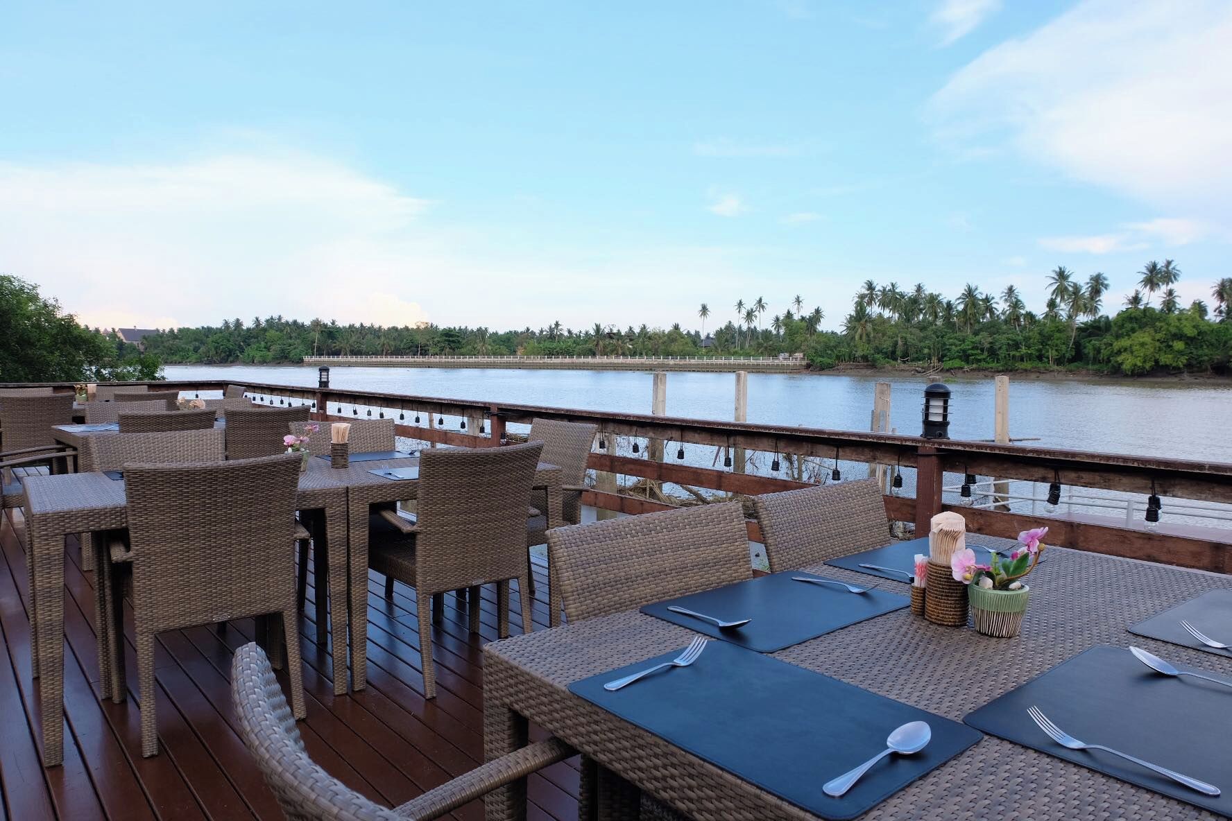 Un espace de restauration en plein air, doté de mobilier et de tables en osier, offrant une vue sur un plan d’eau et des palmiers.
