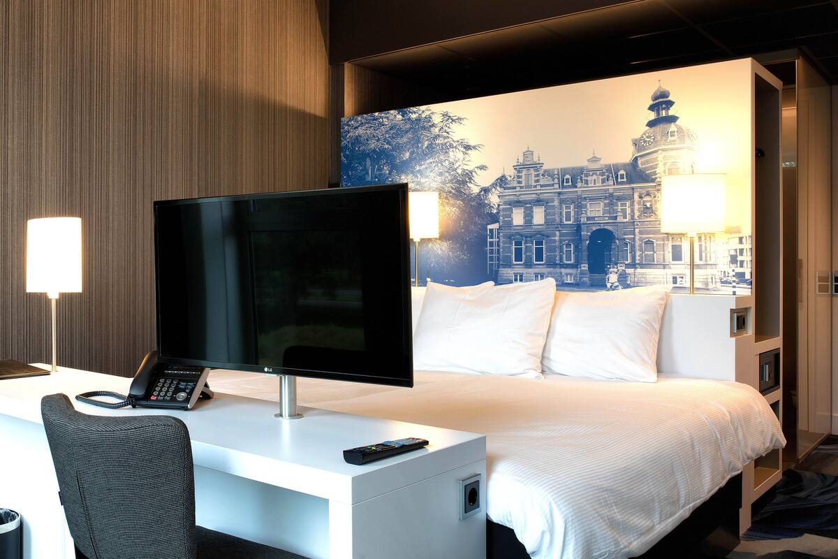 A hotel room with a bed, a desk with a computer monitor, a telephone, and a lamp.