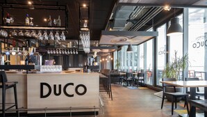 A bar area with a counter labeled "DUCO," shelves with bottles and glasses, and seating arrangements. - Fletcher Hotel - Restaurant Oss (Oss)
