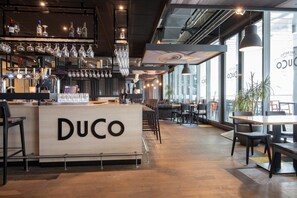 A bar area with a counter labeled "DUCO," shelves with bottles and glasses, and seating arrangements. - Fletcher Hotel - Restaurant Oss (Oss)