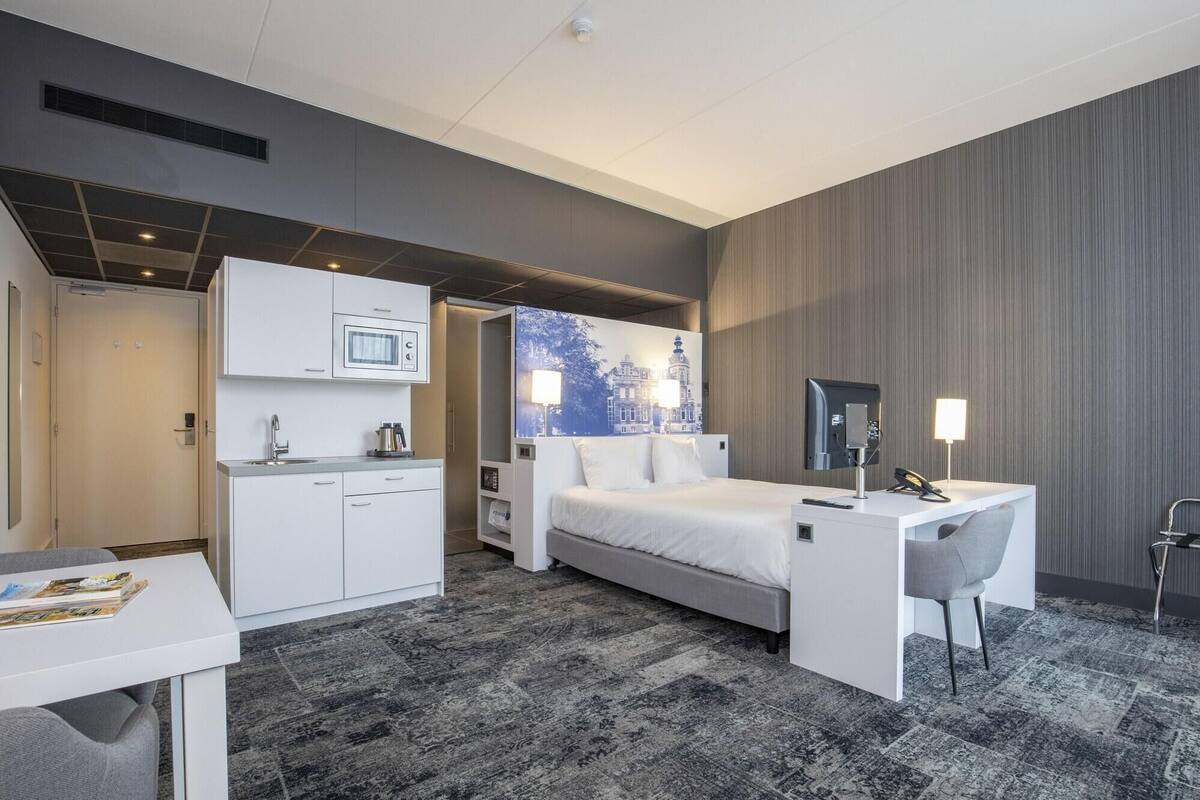 A hotel room with a bed, a desk, a chair, a microwave, and a painting on the wall.