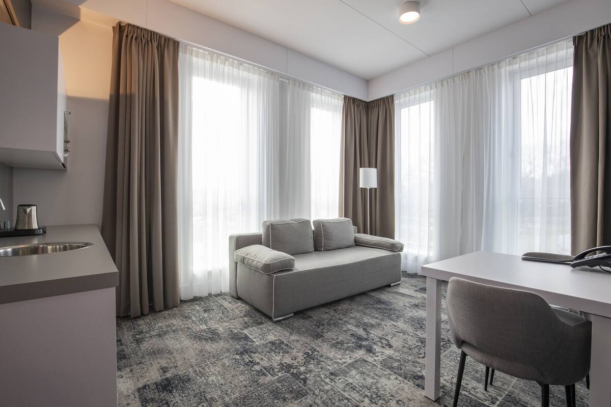 A modern hotel room with a sofa, a dining table, a lamp, and a kitchen area with a sink.