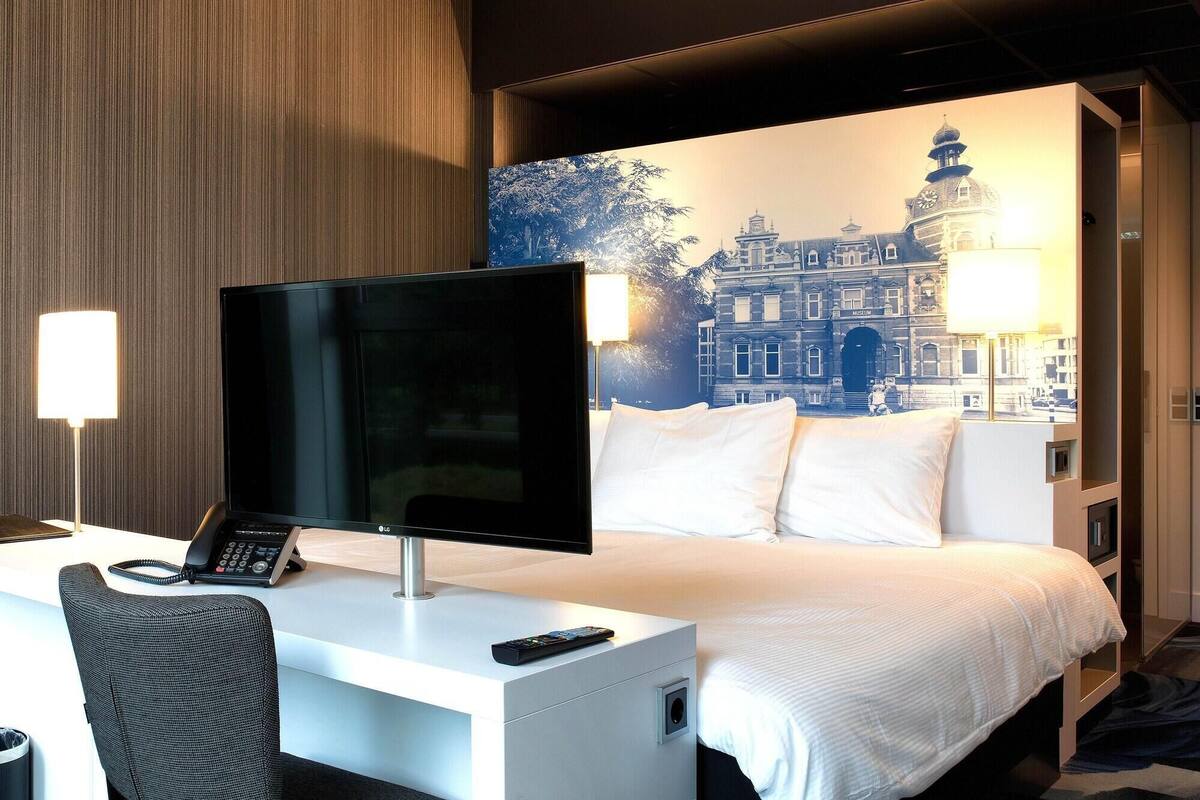 A hotel room with a bed, a desk with a computer and phone, and a large wall mural of a building.