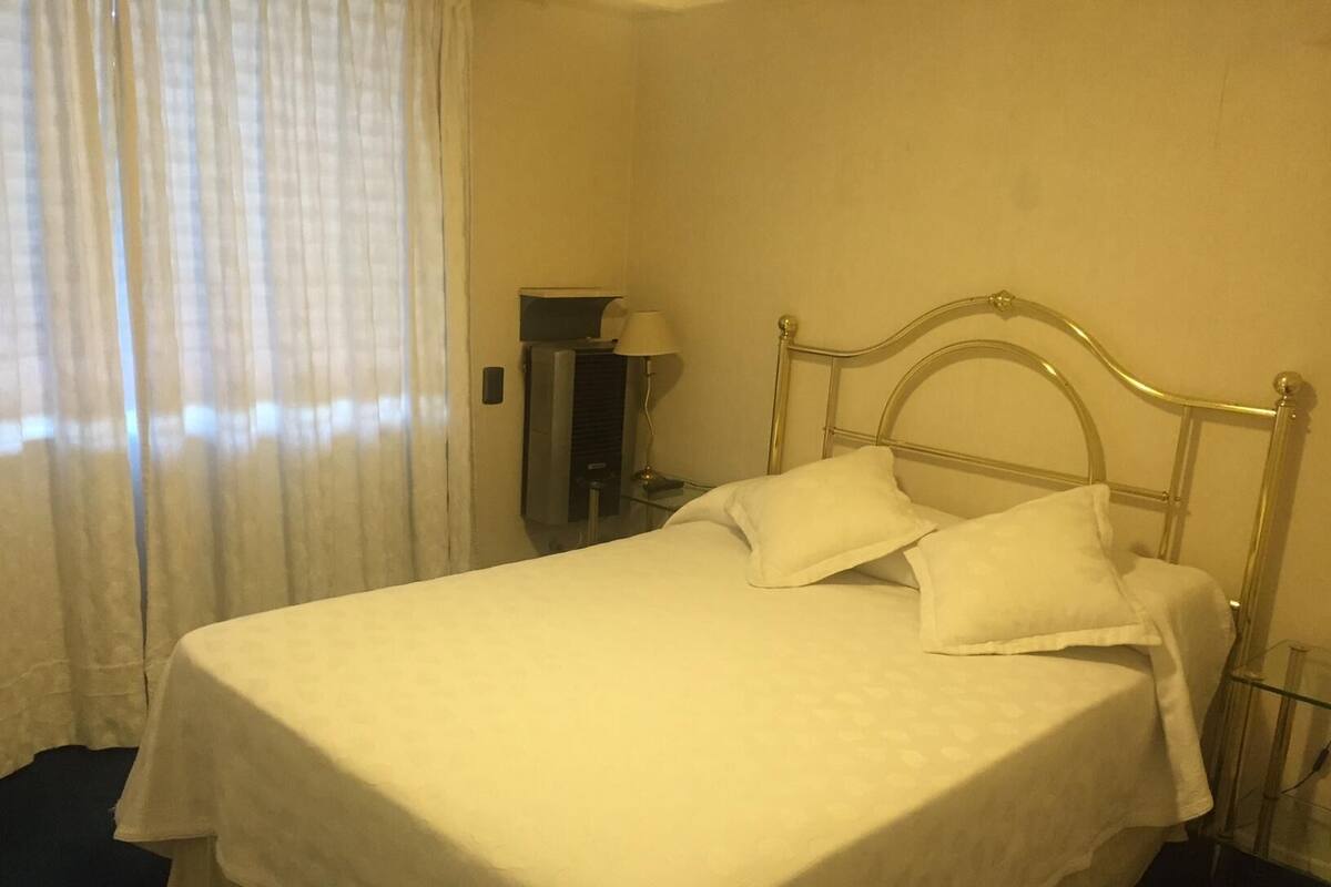 Cama bem arrumada com roupa de cama branca em quarto com parede lisa e janela com cortinas translúcidas.