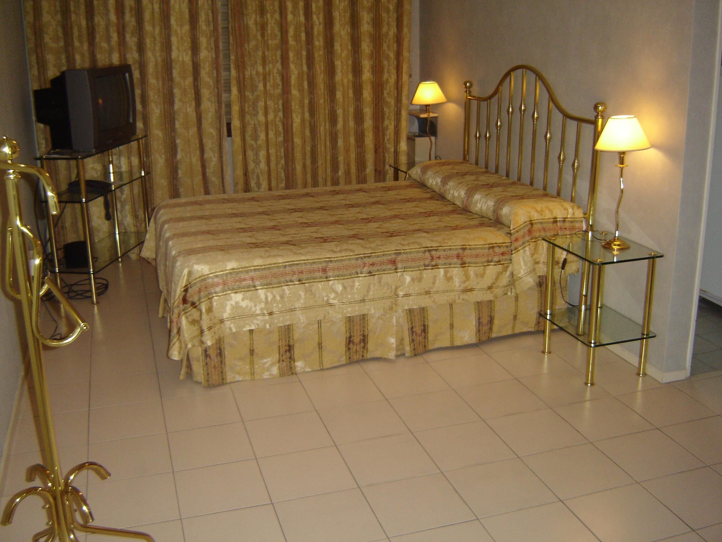 Ein Hotelzimmer mit einem großen Bett, zwei Nachttischlampen, einem Fernseher auf einem Schrank und einem dekorativen goldfarbenen Sessel.