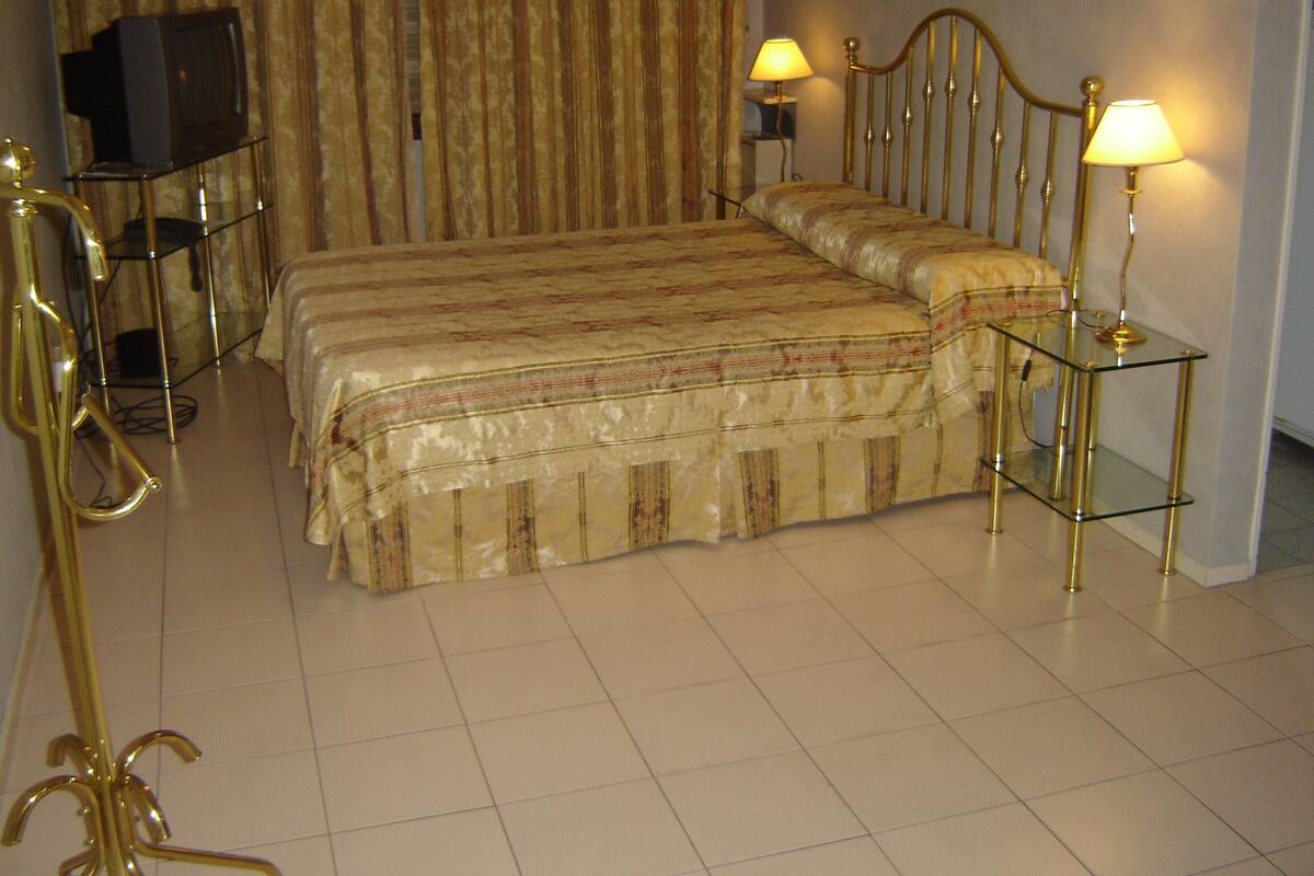 Quarto de hotel com uma cama grande, duas luminárias de cabeceira, uma televisão sobre suporte e uma poltrona dourada decorativa.