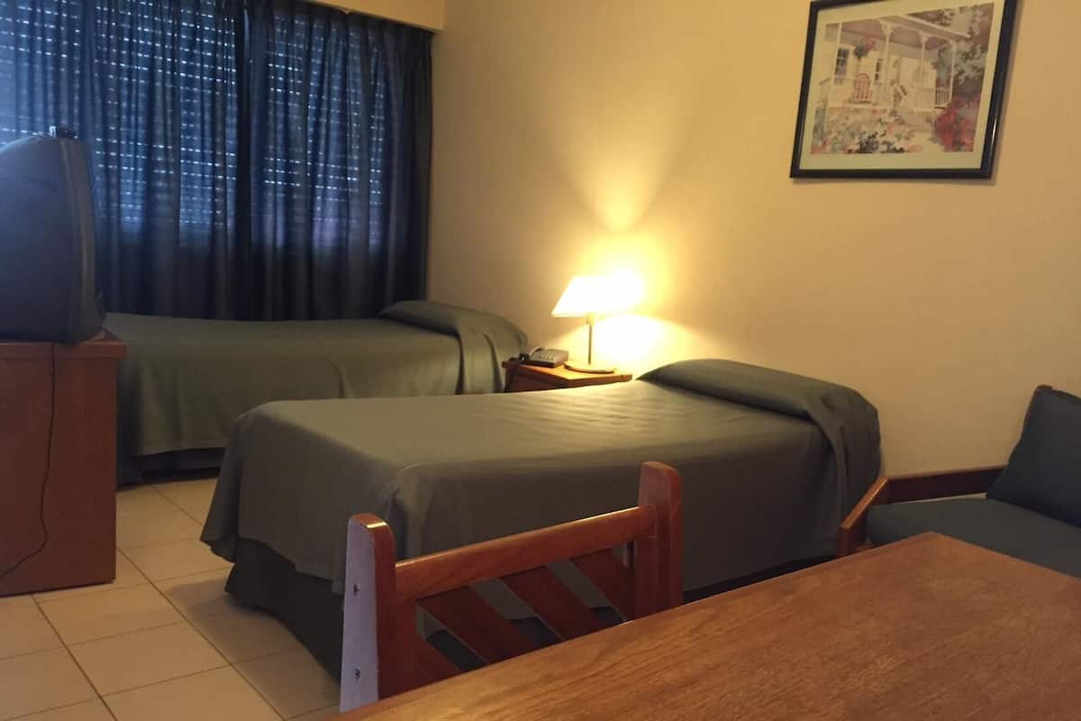 Quarto de hotel com duas camas, uma escrivaninha, uma cadeira, um abajur e um quadro na parede.