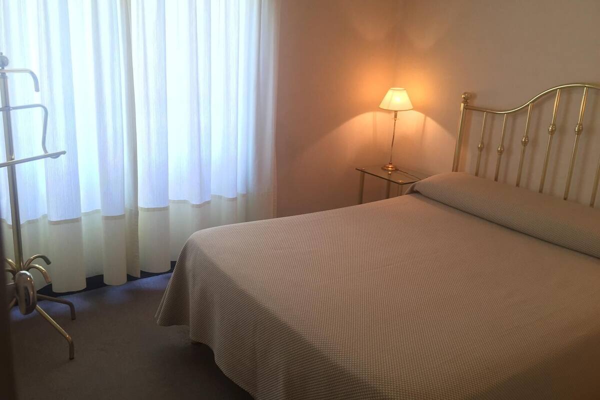 Quarto de hotel com uma cama, mesa de cabeceira com abajur, uma cadeira e janela com cortinas.