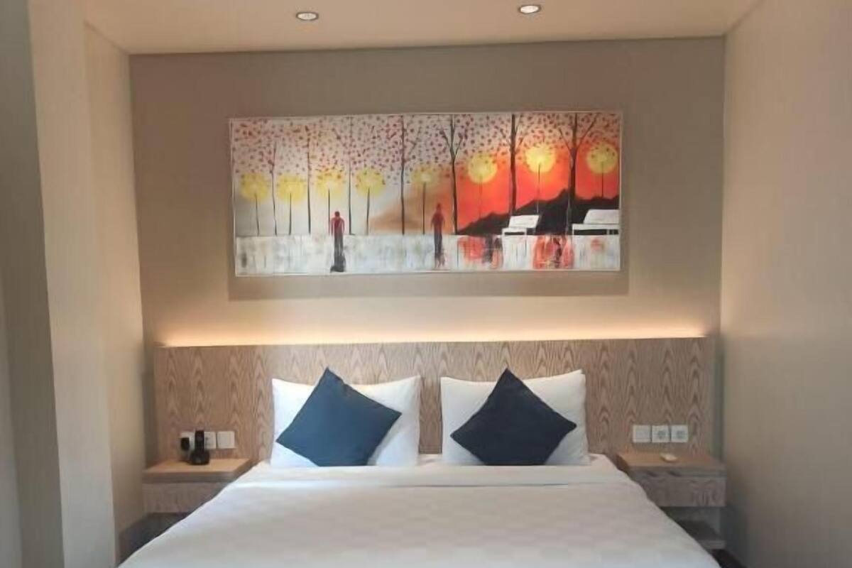 Una habitación de hotel con una cama grande, dos mesitas de noche, un cuadro sobre la cama y suelo de madera.