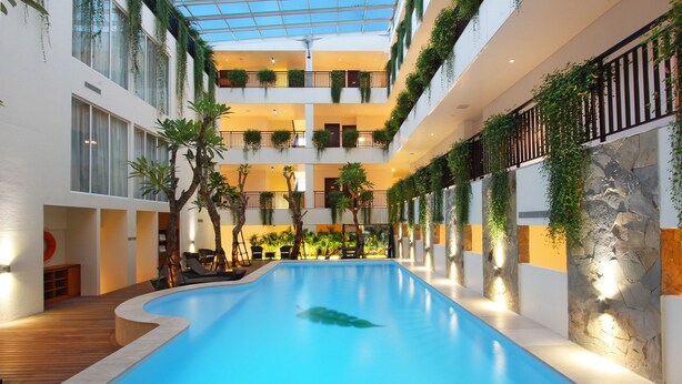 A multi-level indoor swimming pool with a glass ceiling, surrounded by balconies with greenery, and a stone wall on one side. - Dwijaya House of Pakubuwono (Jakarta)
