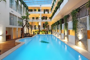 Une piscine intérieure à plusieurs niveaux, dotée d’un toit en verre, entourée de balcons ornés de verdure et bordée d’un mur en pierre d’un côté.