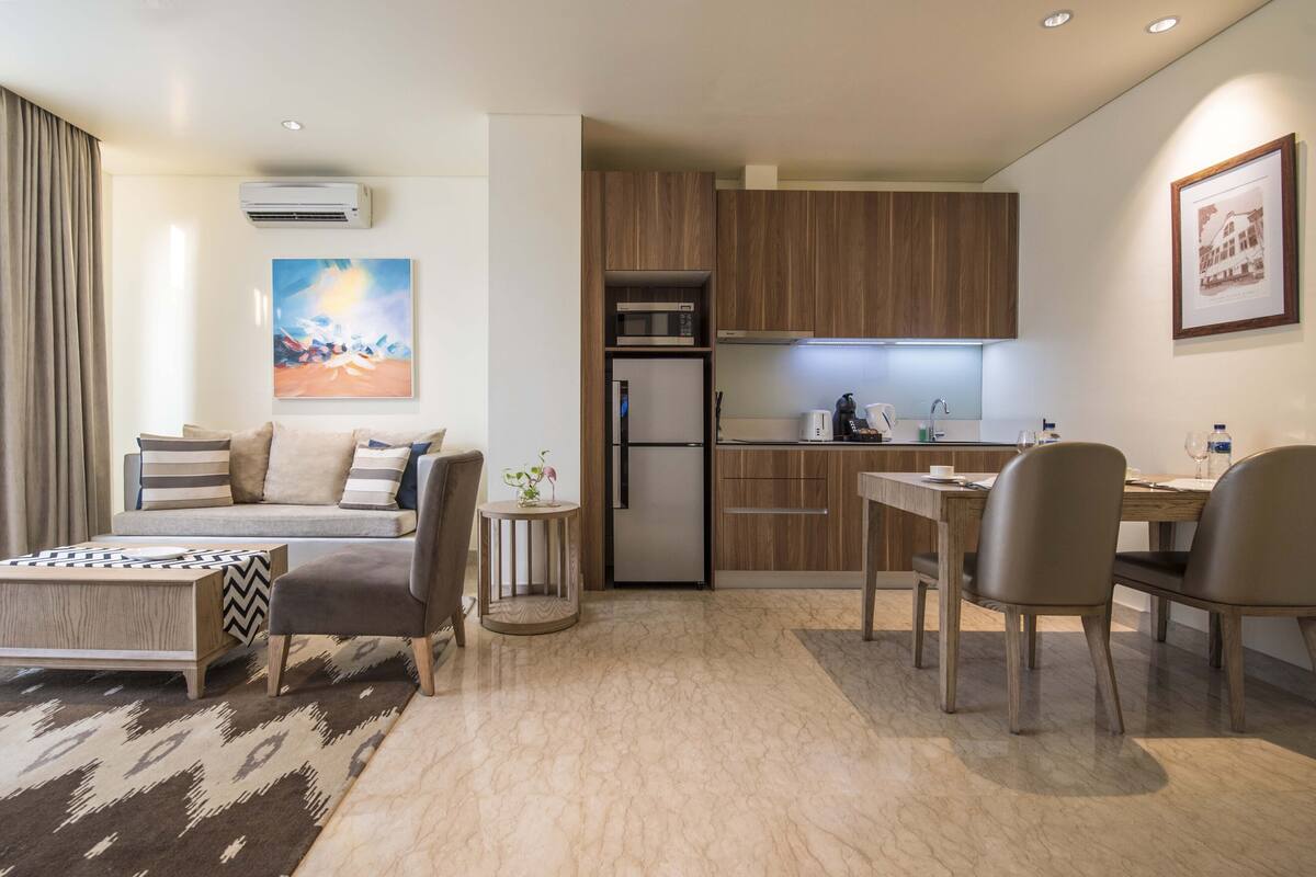 Una sala de estar moderna con un sofá, una mesa de centro y un comedor. Se observa una cocina con armarios de madera y un refrigerador.