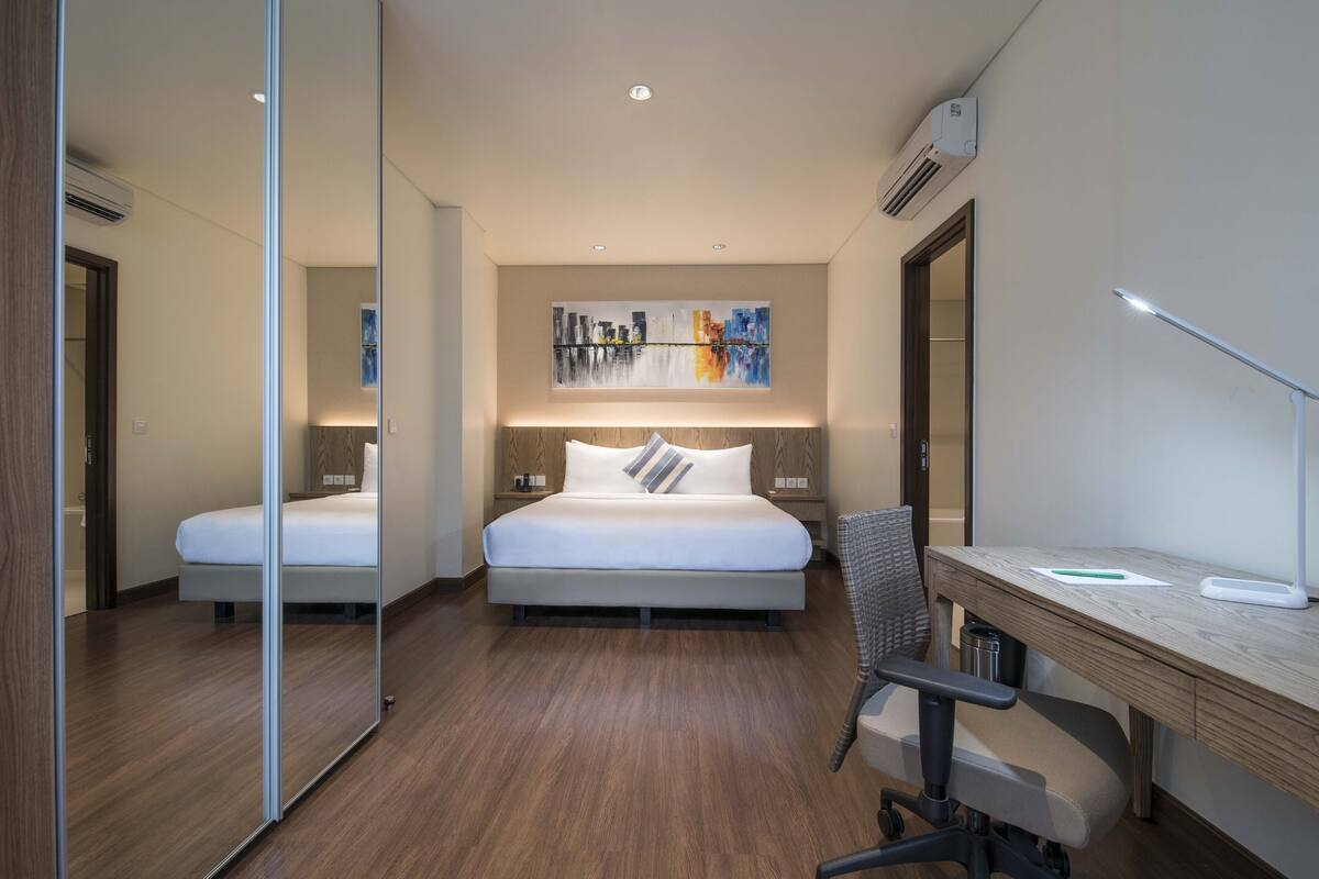 Habitación de hotel moderna con una cama grande, un escritorio y una silla. Hay un cuadro en la pared y un equipo de aire acondicionado sobre la cama.