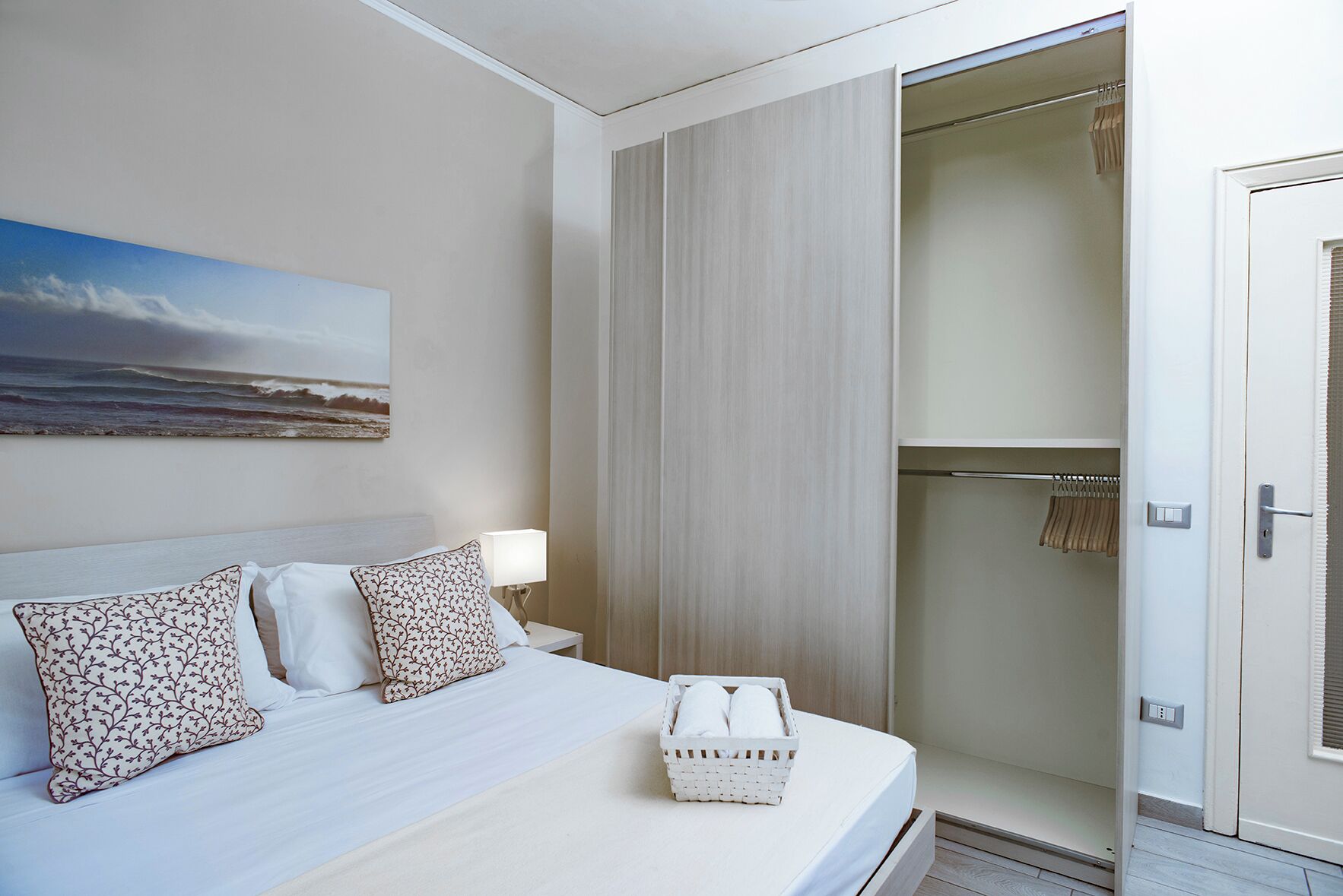 A hotel room with a bed, a nightstand, a lamp, a wall-mounted painting, a closet, and a door.