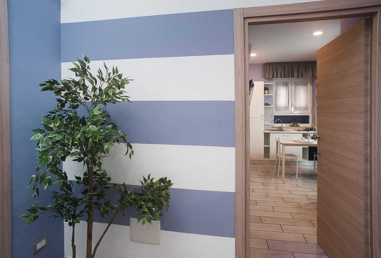 A hallway with a striped wall, a potted plant, and a view into a kitchen area with a dining table and chairs.