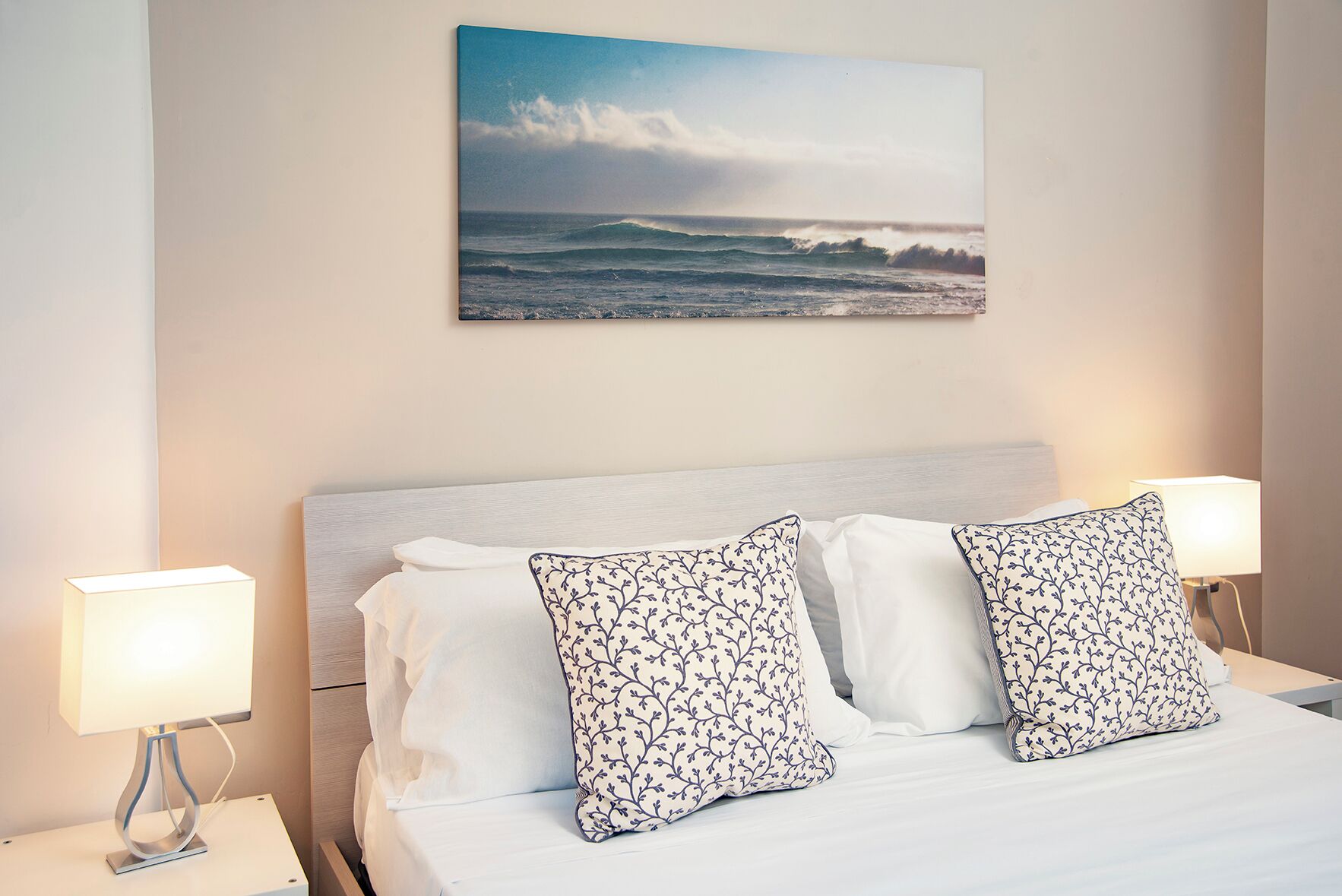 Una cama bien tendida con sábanas blancas y dos almohadones decorativos, un cuadro colgado en la pared que muestra una escena de playa y lámparas de noche a cada lado.