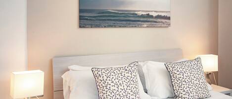 Una cama bien tendida con sábanas blancas y dos almohadones decorativos, un cuadro colgado en la pared que muestra una escena de playa y lámparas de noche a cada lado.