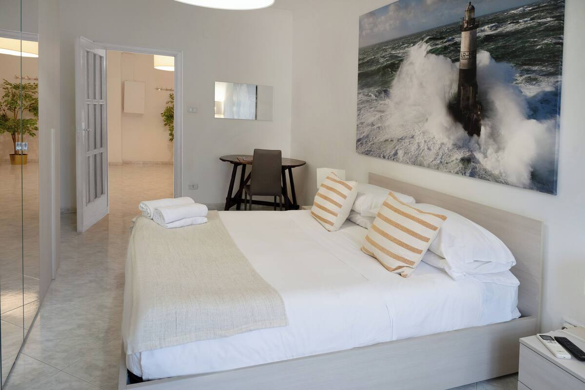 Un dormitorio con cama, mesita de noche, silla, un cuadro de un faro y vistas a un pasillo.