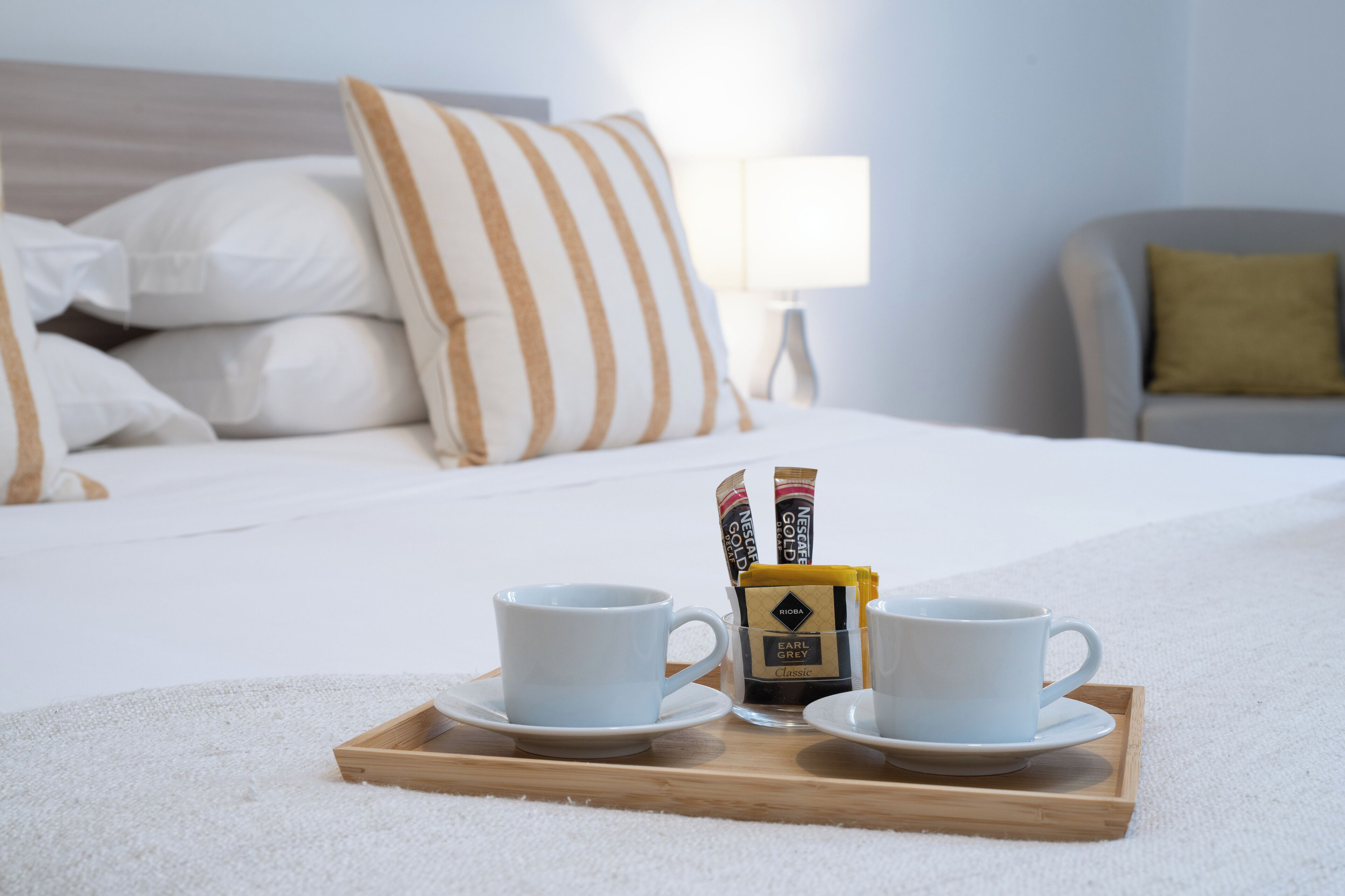 Una cama bien hecha con almohadillas blancas y a rayas, una mesita de noche con una lámpara y una bandeja con dos tazas y un paquete de café.