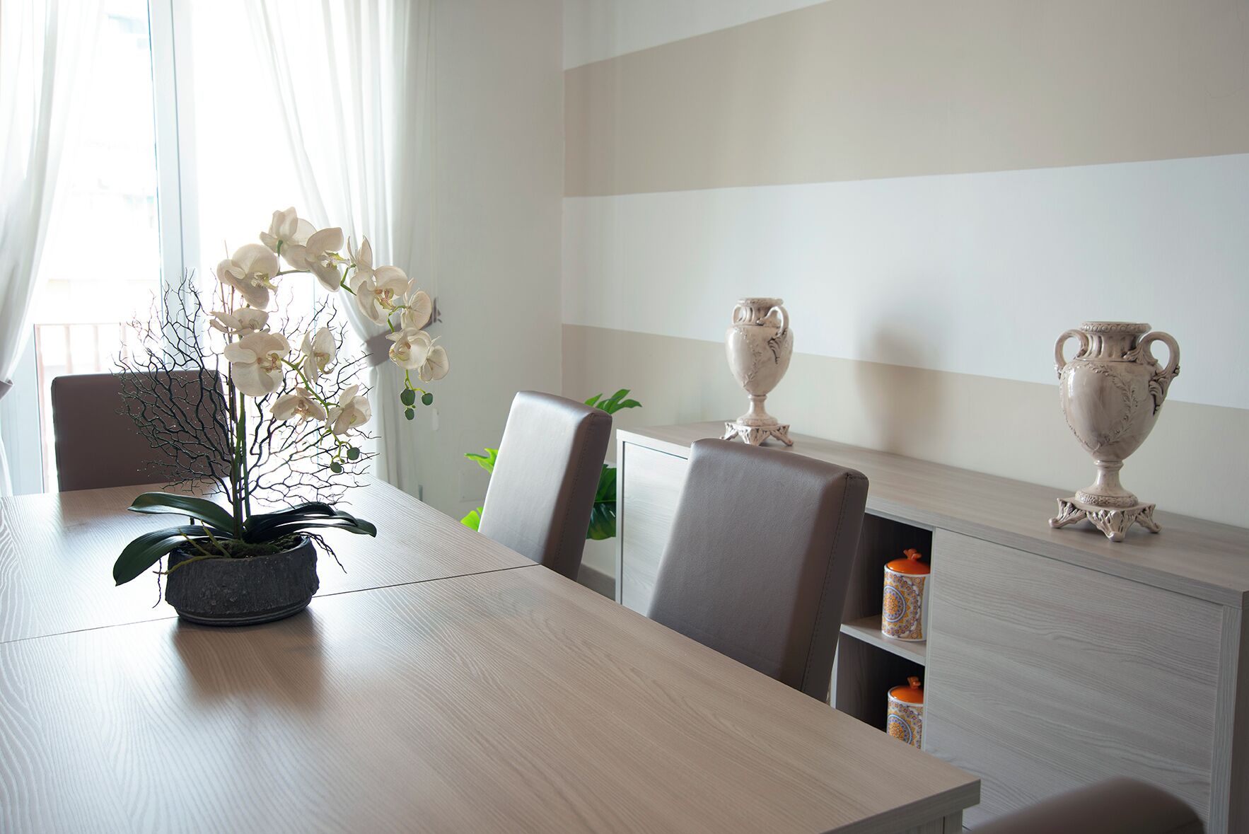 A modern dining area with a wooden table, two chairs, a potted orchid, and two decorative vases on a shelf.
