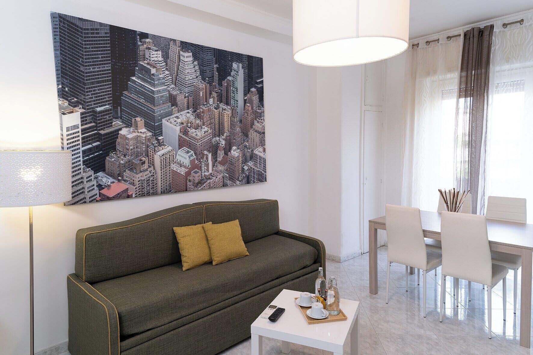 Ein modernes Wohnzimmer mit einem grünen Sofa, einem Couchtisch und einem Essbereich mit weißem Tisch und Stühlen. An der Wand hängt ein großes Stadtbild.