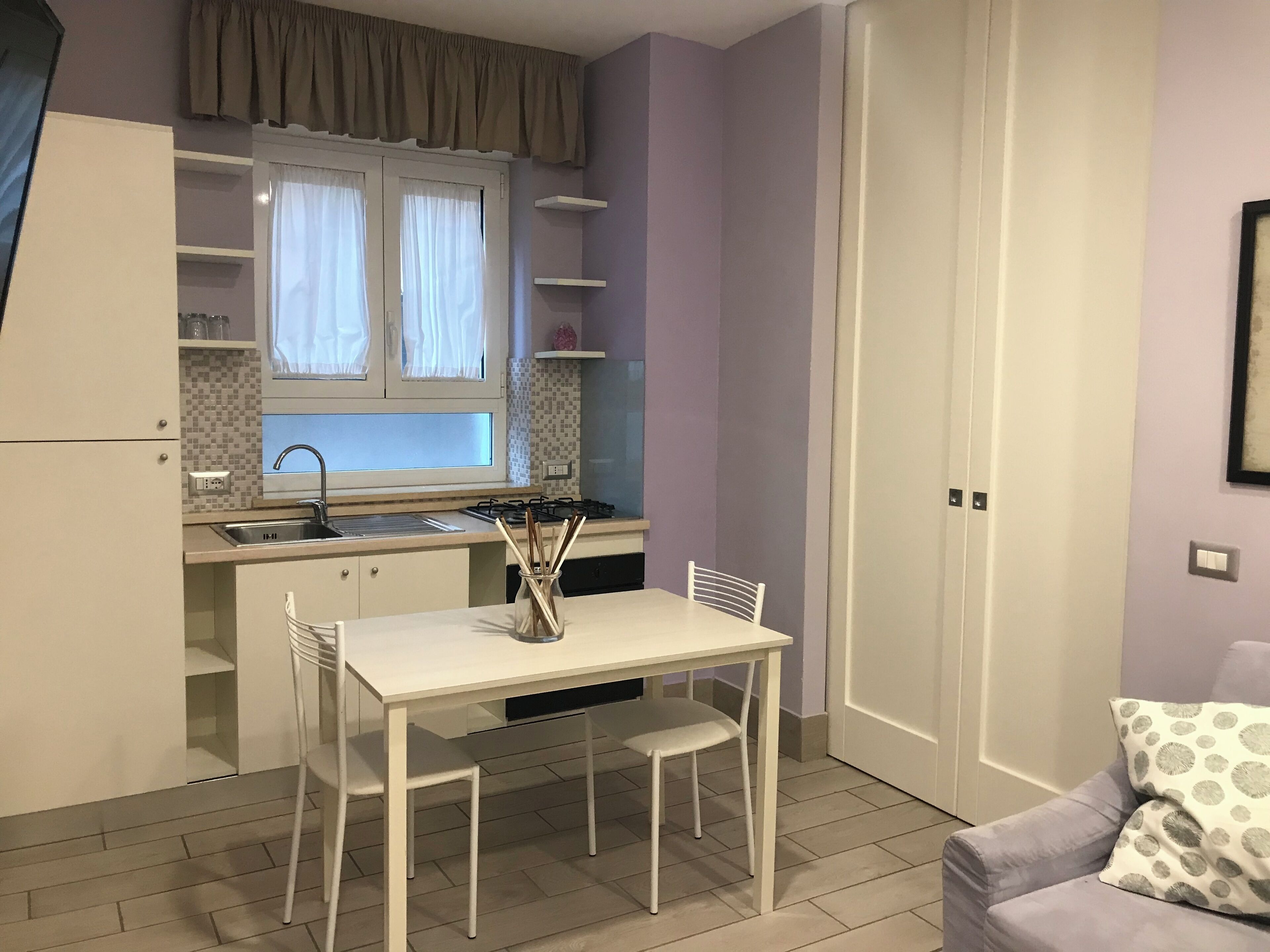 A compact kitchen with white cabinets, a window with curtains, a sink, and a dining area with a table and chairs.