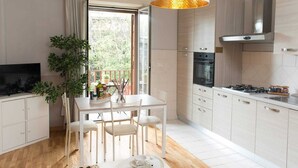 A modern kitchen with white cabinetry, a dining table with chairs, a TV, and a large window with a view of greenery. - Residenza Flegrea (Naples)